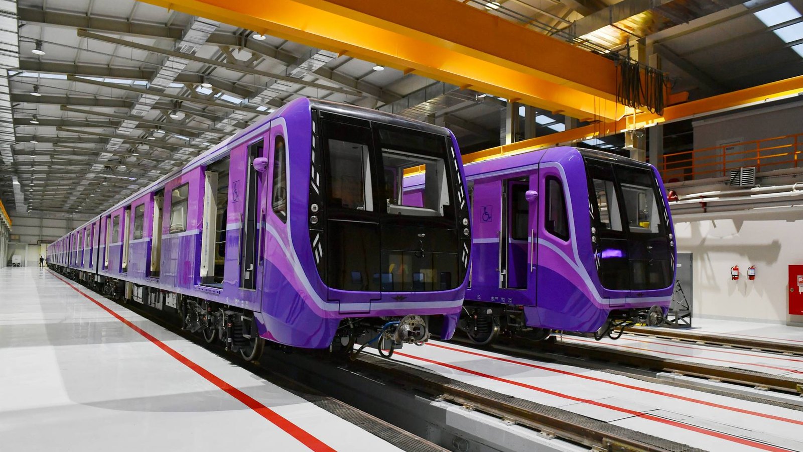 Two purple metro trains on tracks inside a bright depot hall with yellow overhead cranes