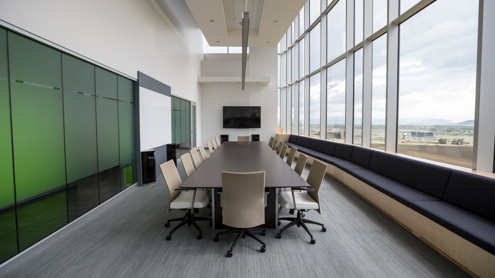 Long conference table with beige chairs in a bright room with floor-to-ceiling windows and a wall screen