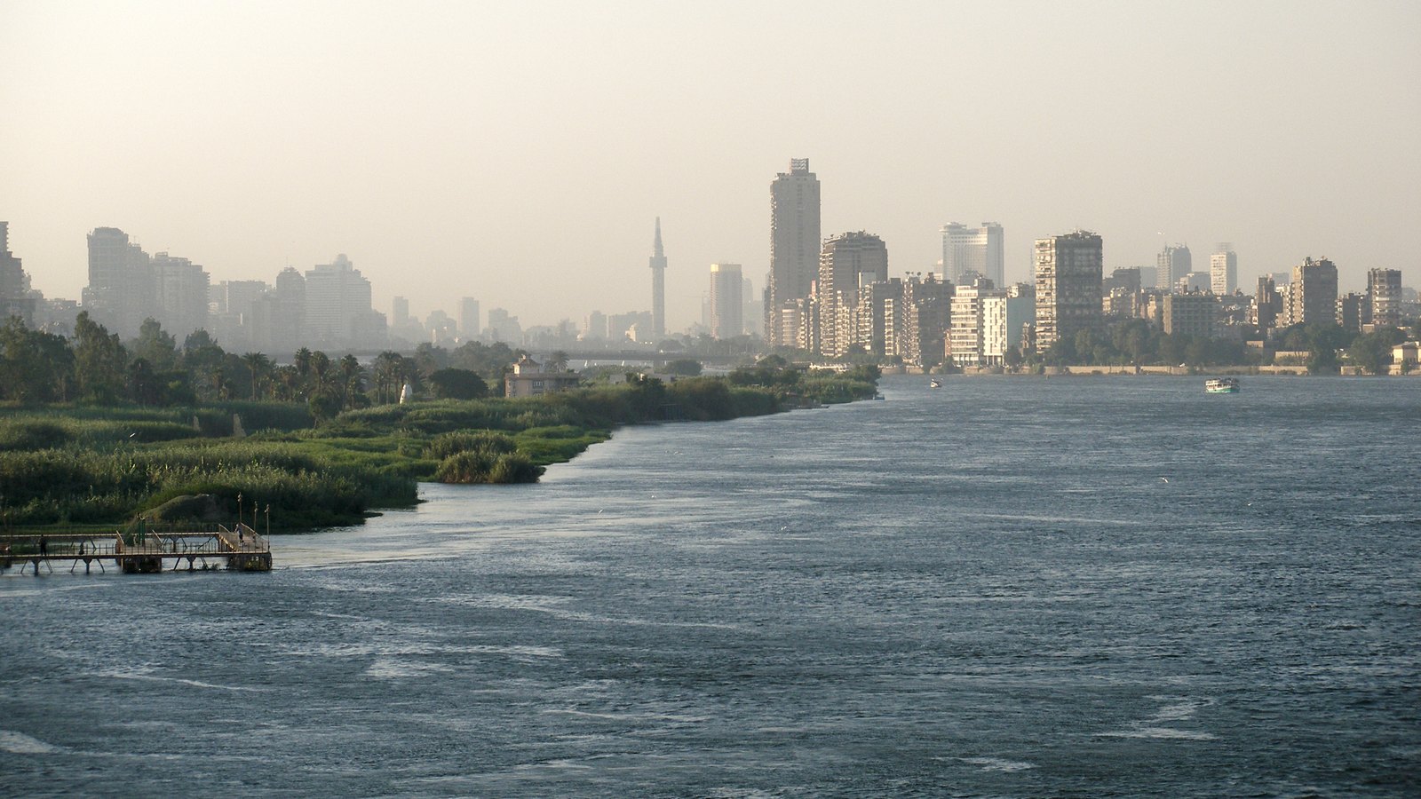 Hazy city skyline with a slender tower and high-rise blocks beside a wide stretch of the Nile