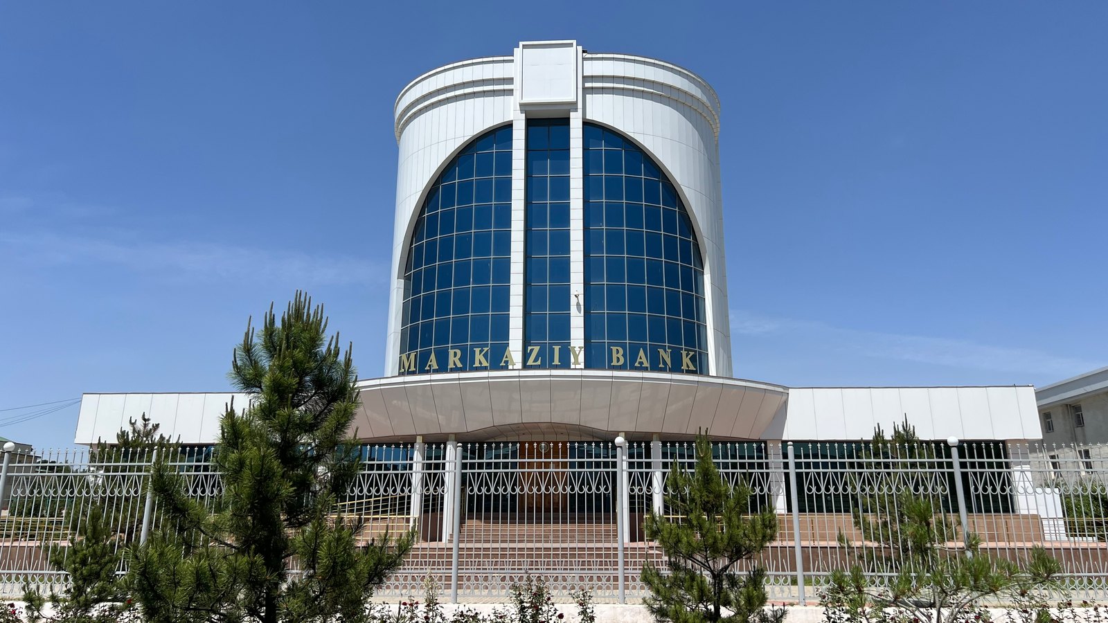 Rounded blue glass tower of a central bank branch with Markaziy Bank lettering, behind a fence and young pines