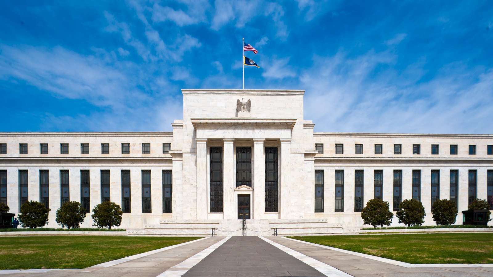 White marble neoclassical facade of the Federal Reserve headquarters under a blue sky with streaky clouds