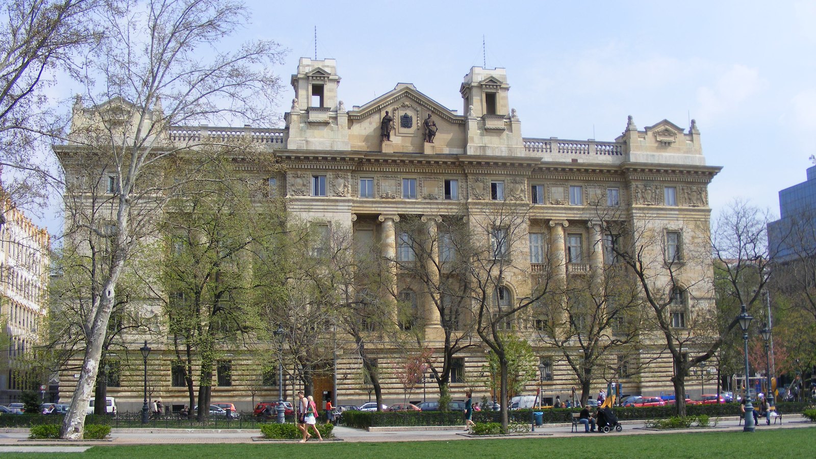 Ornate stone bank building with columns and rooftop sculptures behind a green lawn and bare spring trees