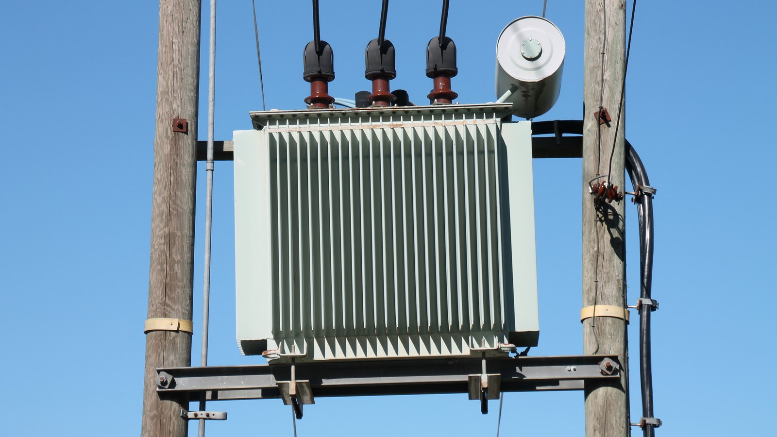 A grey finned transformer mounted between two wooden poles, with three insulated cable connections on top