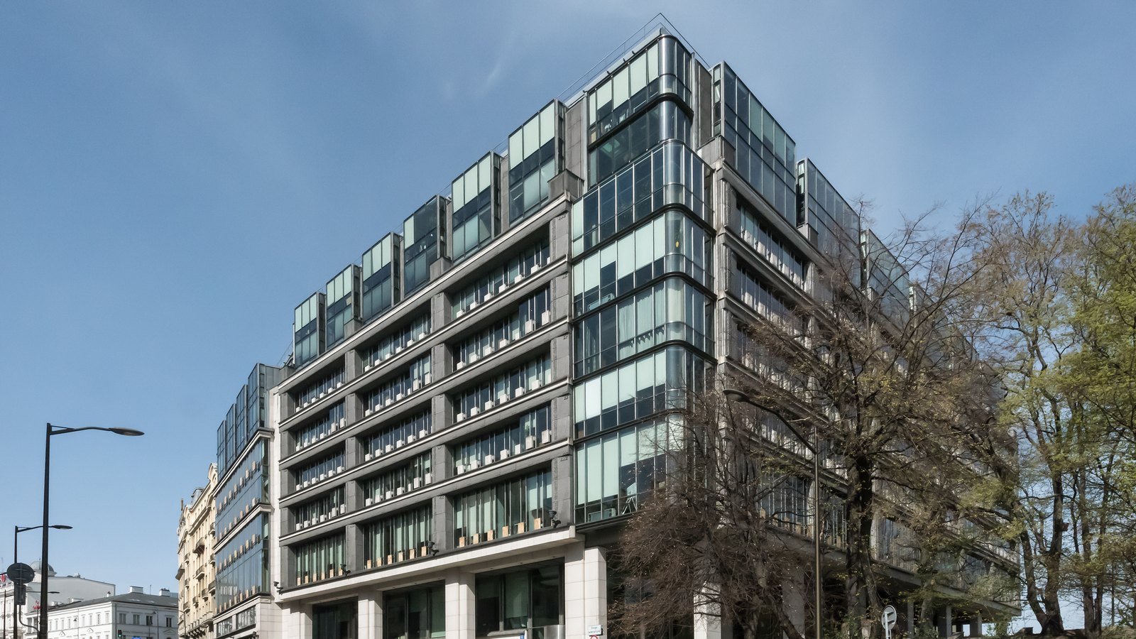 A modern glass and steel office block on a sloping street corner, with bare trees beside it