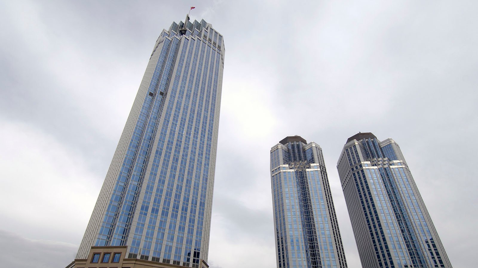 Three glass skyscrapers, the tallest topped by a flag, rise above a low stone building under a grey sky