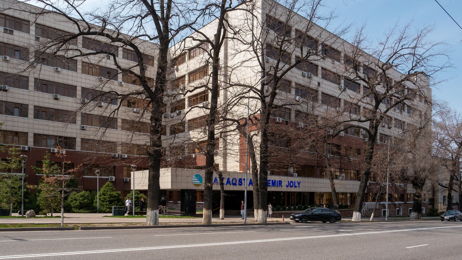 A tall office block with a sign reading Qazaqstan Temir Joly, behind bare trees on a wide street