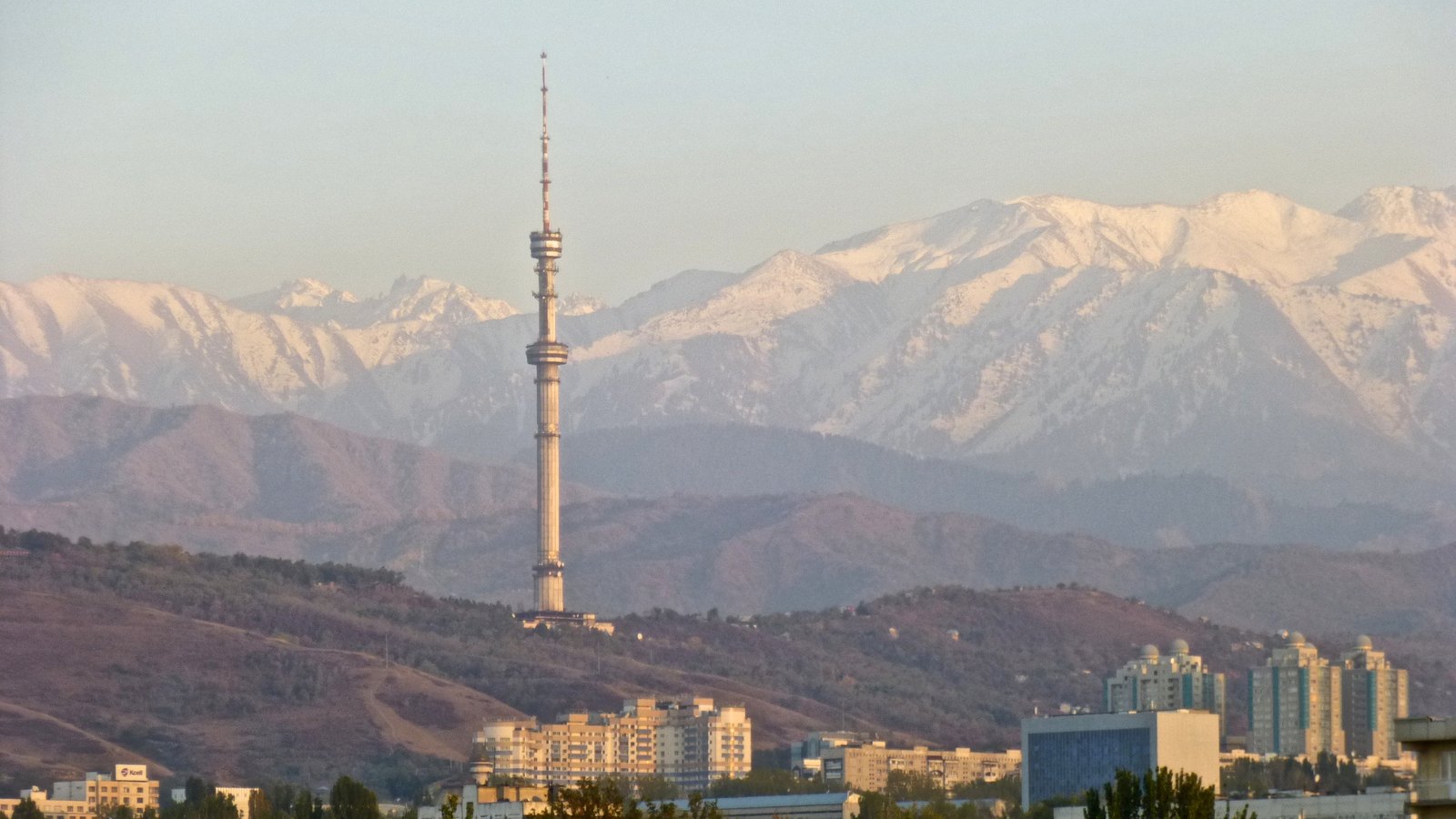 A slender television tower on a wooded hill in front of snow-capped mountains, with apartment blocks below
