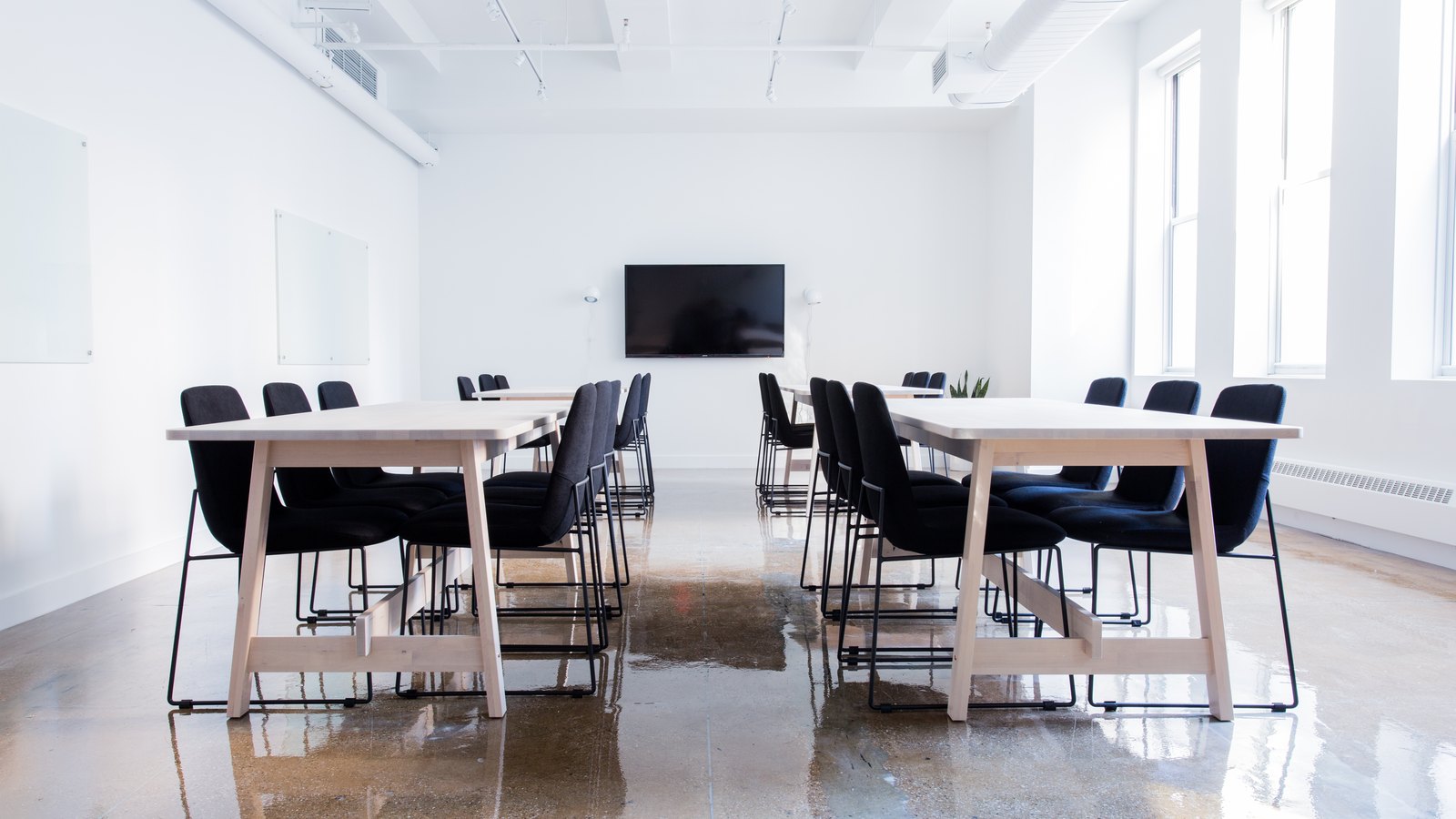 Two long tables with black chairs in a bright white boardroom with tall windows and a wall screen