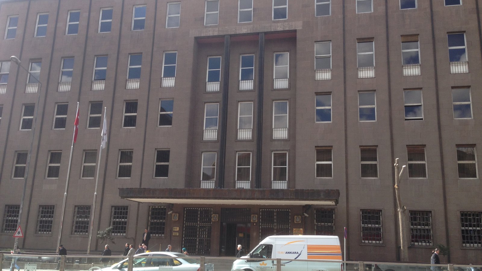 A long brown stone office building with rows of windows and a canopy over the entrance, cars passing in front