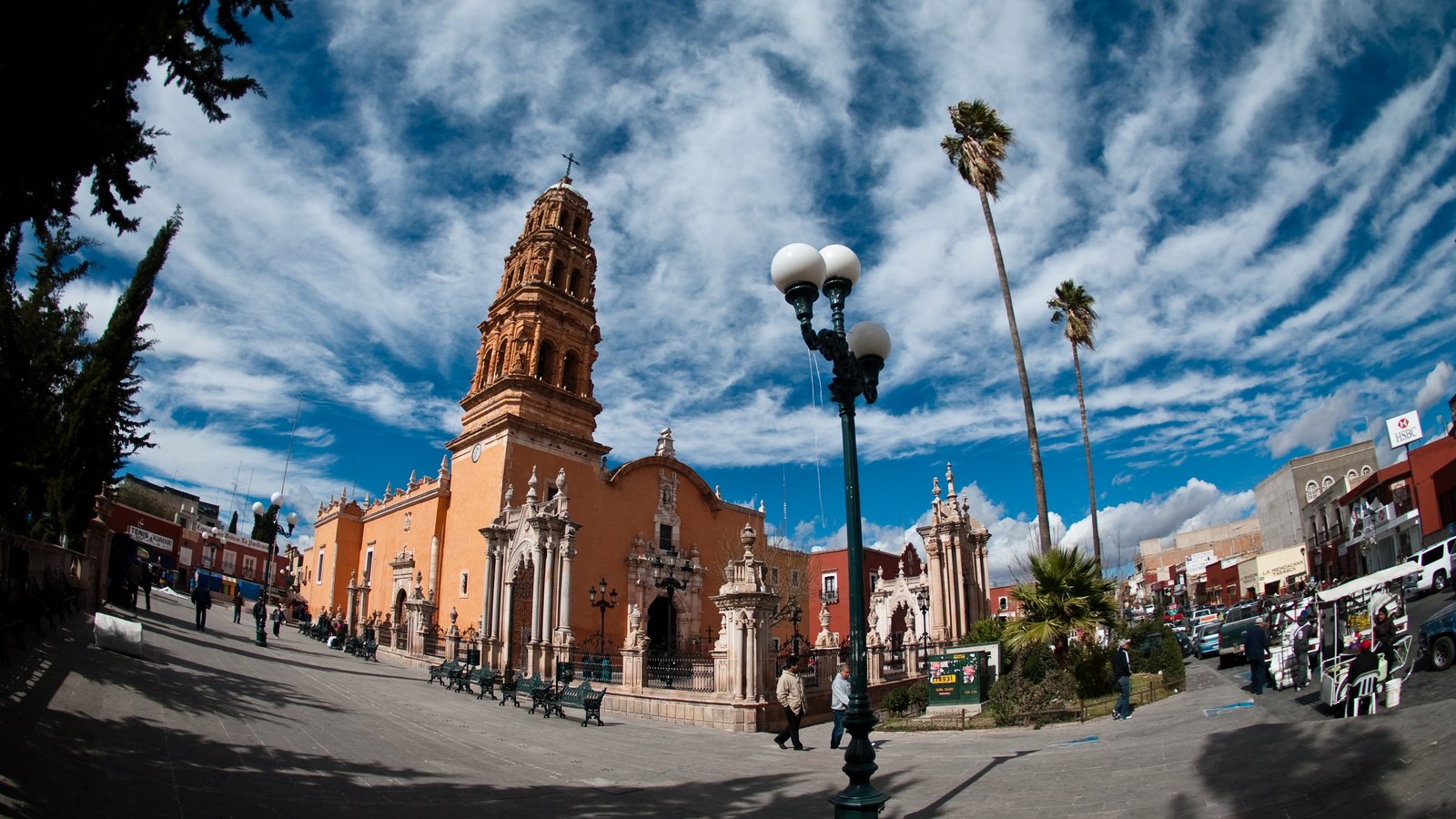 An orange baroque church with a bell tower on a square with palm trees and lamps under a cloudy sky