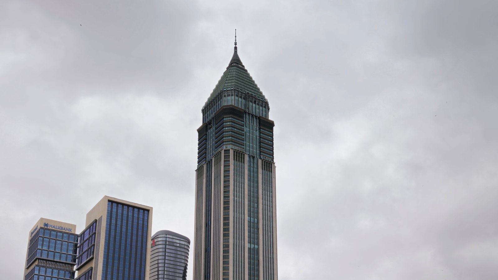 A pointed skyscraper rising among dark glass office towers under a cloudy sky