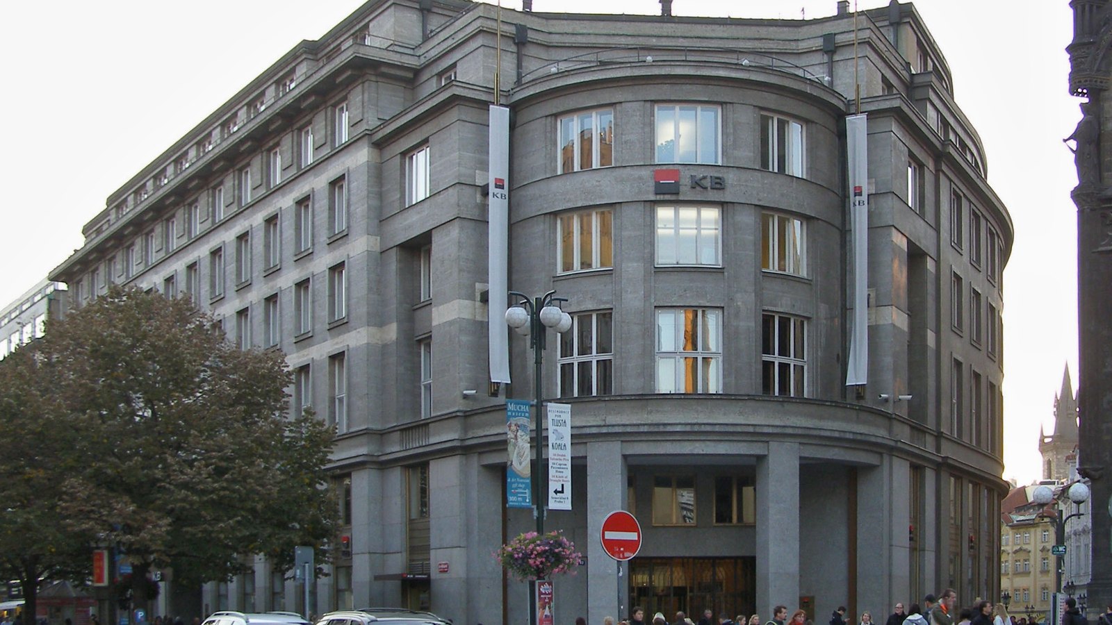 A grey stone bank building with a KB sign on its rounded corner, above a busy pavement