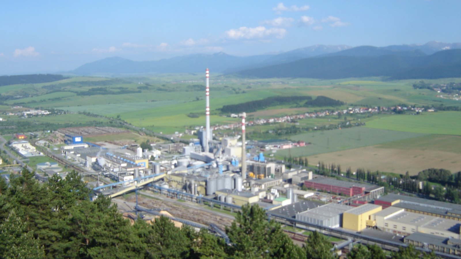 A large paper mill with chimneys spread along a valley below wooded hills