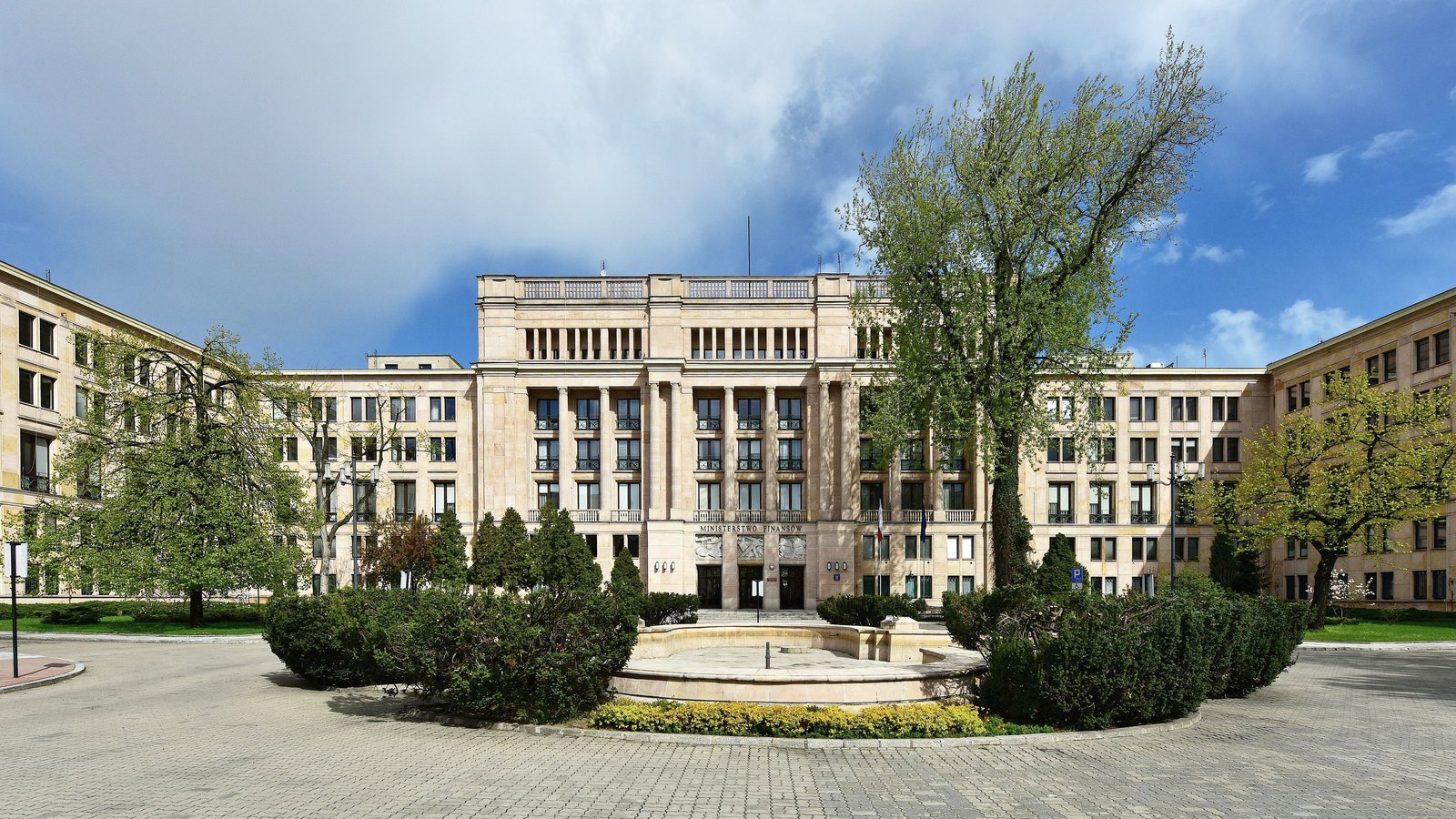 A large stone ministry building with tall pilasters behind a round garden and fountain