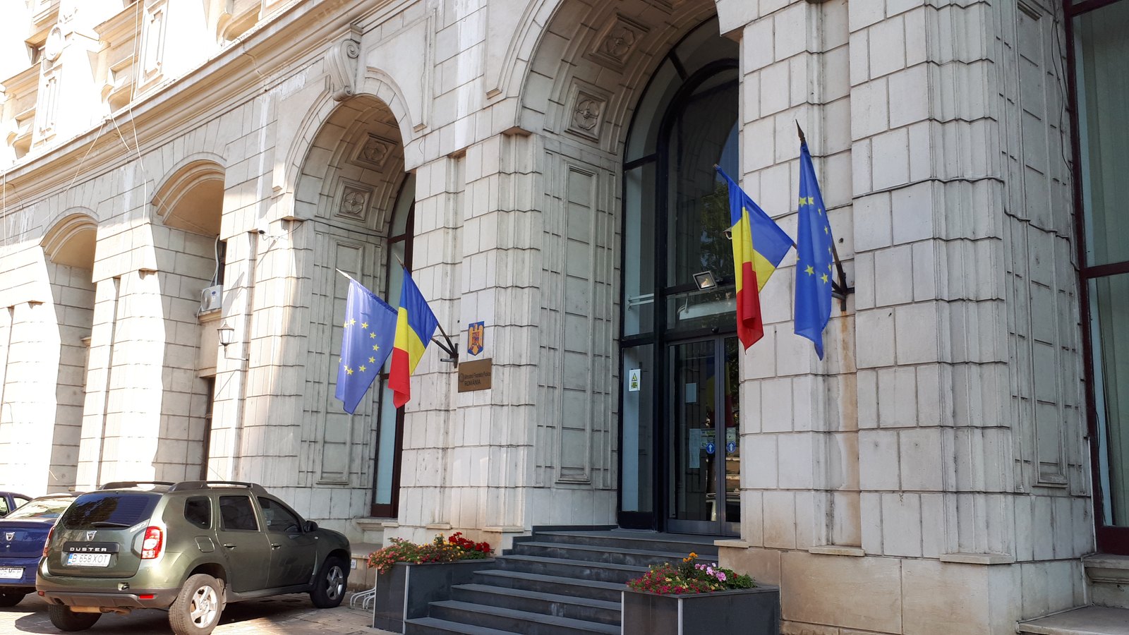 The stone entrance of a ministry building with Romanian and EU flags beside its arched doors