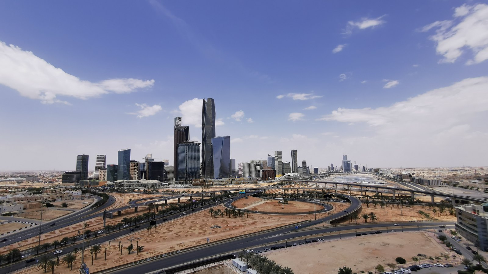 Glass skyscrapers of Riyadh's financial district beyond highways and open ground under a blue sky