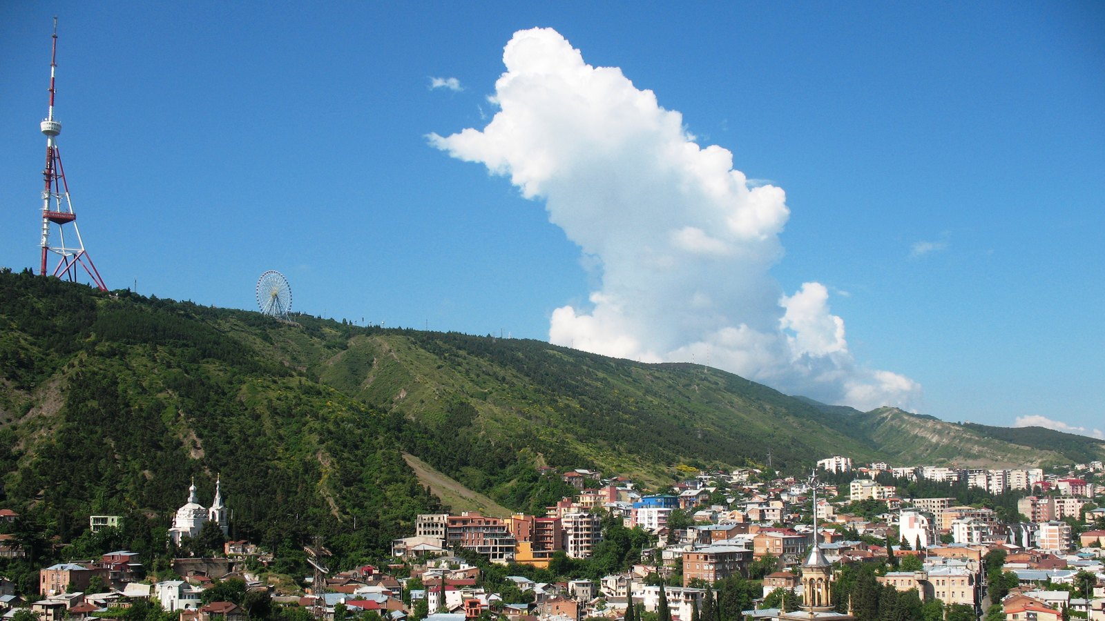 Old Tbilisi rooftops below a green ridge topped by a red and white TV tower, with a tall white cloud above