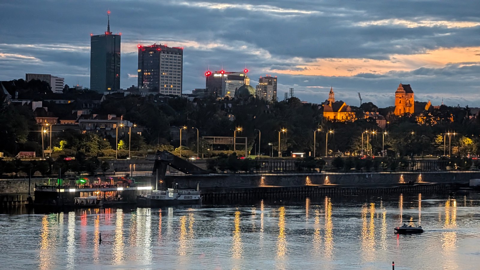 Warsaw high-rises and historic spires at dusk, with city lights reflected in the Vistula river